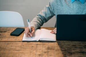 ノートパソコンを置いた木の机でノートに書き込む手元